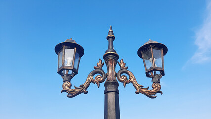 A symmetrical, low-angle photograph of a vintage double-headed street lamp with ornate wrought-iron scrollwork and bronze-gold accents.