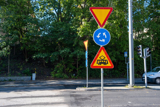 Traffic sign at road roundabout showing warning near intersection on urban street transport route beside green trees in bright daylight
