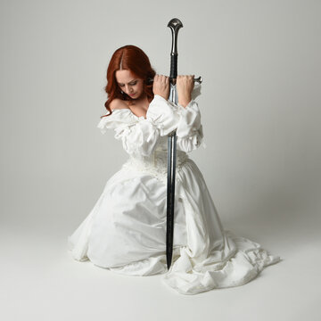 full length portrait beautiful female model wearing historical white fantasy wedding dress, fairytale ball gown  holding sword weapon. isolated figure sitting pose on studio background.