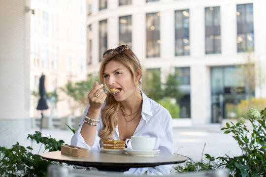 Elegant Female Sitting at a Cafe Table with Layered Cake, Hot Drink, and Urban Greenery Around