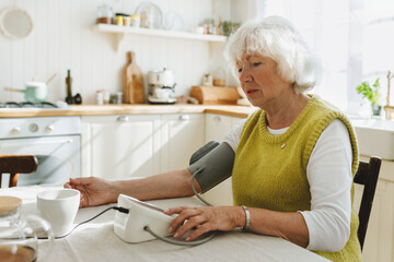 Side view of senior lady measuring blood pressure at home feeling unwell as weather change affection, looking at digital monitor while wearing aneroid cuff on her shoulder sitting at kitchen table