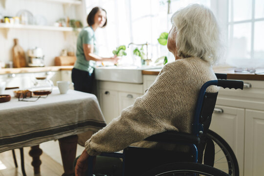 Rare view image with selective focus on old disabled female in wheelchair talking to her home assistant or caregiver cleaning kitchen or cooking dinner standing next to window in front of counter