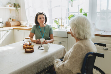Selective focus of caregiver therapist counseling her old female disabled patient with alzheimer in wheelchair both sitting at kitchen table, senior lady pictured with back turned at camera