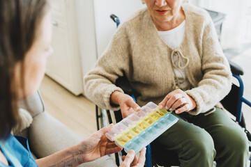 Indoor image of two unrecognizable people, caregiver holding pill box giving medication to senior lady in wheelchair after brain stroke to recover, helping to take remedy safely, at right time