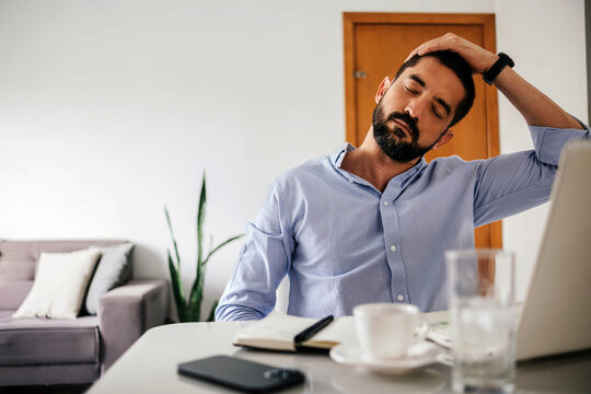 Adult business man stretching his neck while sitting at a desk in a home office during remote work. Concepts of ergonomics, self care, work break, posture, health and productivity