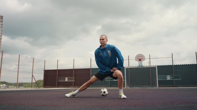 soccer player lunging on urban court with ball, heavy clouds and damp surface, athlete in blue windbreaker performing dynamic lateral stretches and control drills, neon cleats gripping asphalt,