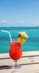 Obraz premium Tall glass with red beverage, straw, ice and pineapple slice with cherry and umbrella against a turquoise sea and blue sky, representing refreshment