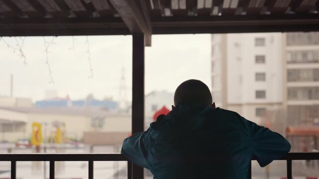 man on balcony watching rain, cradling football and staring at blurred city skyline through droplets, contemplative solitude, muted colors, quiet longing and reflective pause in apartment terrace