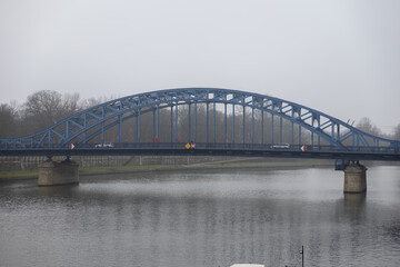 Obraz premium Prominent blue arched truss bridge structure spans a wide urban river, symbolizing critical infrastructure, modern connectivity, and robust engineering under an overcast sky