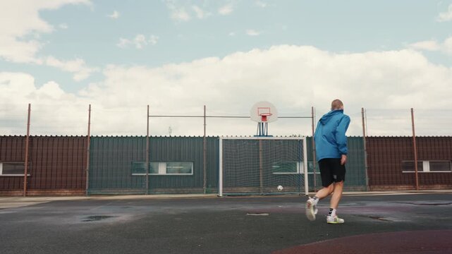 Caucasian player on wet urban court, blue jacket juggling ball toward goal, puddles reflecting cloudy skyline, fence lined rooftop arena, warming up with focused footwork, running to shoot