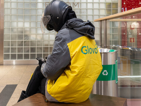 Krakow, Poland - December 2, 2025: Glovo worker in bright uniform, wearing helmet, sits checking smart phone, showing urban gig economy, fast delivery job