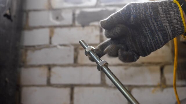 Close-up of a construction worker's hands in protective gloves meticulously twisting and tightening a hex nut onto a long metal threaded rod against a white brick wall background
