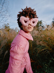 Obraz premium Child wearing a pink outfit and fluffy pink mask in an outdoor meadow, posing with curiosity among tall grasses and a bright sky, conveying imaginative dress up and playful fashion in nature