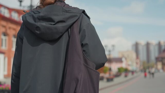 redhead person listening to music outdoors amid city buildings, wearing black jacket and backpack, large headphones, looking up at blue sky with soft clouds, reflective and calm mood
