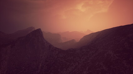Obraz premium Layered mountain peaks create a stunning backdrop as a warm, dreamy sunset casts an enchanting glow. Clouds drift slowly, adding to the mystical atmosphere of the landscape.