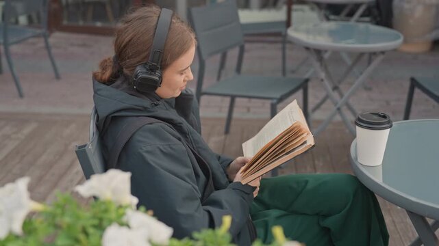 Woman reading at outdoor caf wearing headphones, bundled in dark jacket, sipping coffee from togo cup while turning pages of paperback on wooden patio table surrounded by white flowers and potted