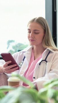 Vertical video: Opening shot provider in lab coat sipping coffee scrolling phone in lounge on break