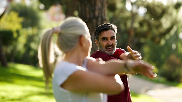 Mature man and woman doing arm stretching exercises together outdoors in a sunny park, actively embracing a healthy lifestyle