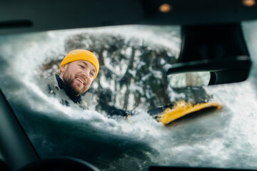 Smiling man scraping ice off car windshield in winter