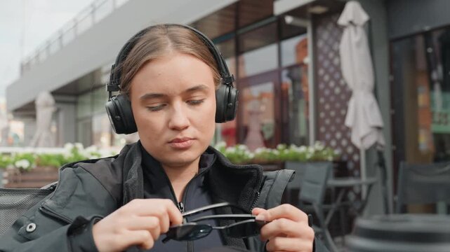 caucasian woman at cafe wearing headphones, rummaging through backpack, adjusting sunglasses, sipping coffee from takeaway cup, braided hair visible, focused expression, urban terrace setting