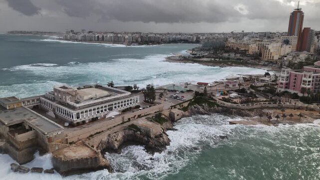 Aerial view of the turquoise sea crashing against the shore near buildings and hotels under a cloudy sky, St. Julian's, Malta.