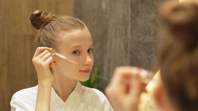Cute Teen Girl Applying Facial Serum With Dropper in Modern Bathroom, Looking in Mirror. Blonde Caucasian Gen Alpha Girl and Beauty Routine. Daily Skincare Routine