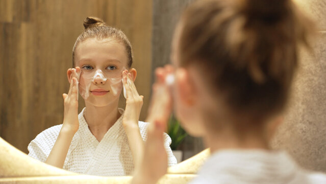 Cute Smiling Teen Girl Applies Cleansing Foam to Face in Front Mirror in Modern Bathroom. Blonde Caucasian Gen Alpha Girl Washing Face. Skincare routine for youthful, healthy skin. Beauty Routine.