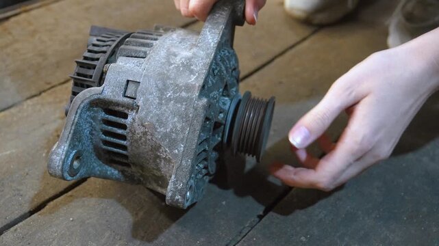 Female hands inspecting and spinning the pulley of a used automotive alternator on a wooden floor, checking the condition of the bearing and searching for signs of wear and tear