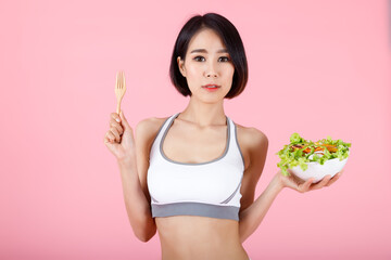 Beautiful Asian woman in sportswear holding fresh vegetable salad bowl and wooden fork. Healthy girl ready to eat clean food for diet concept isolated on pink background.