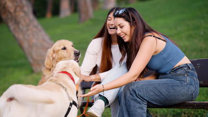 Two cheerful women laughing while interacting with a happy golden retriever and a labrador dog on a bench in a green park