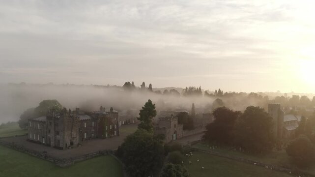 Aerial view of Ripley Castle and grounds partially obscured by a dense fog, with warm sunlight filtering through the mist, Ripley, England, United Kingdom.