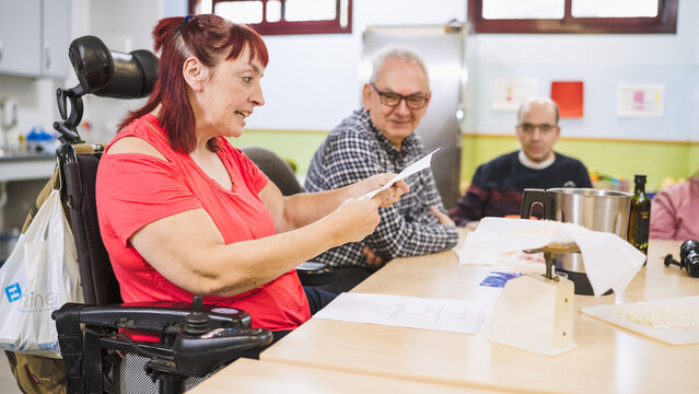 Woman with disability sitting in a wheelchair and reading a recipe in a cooking class with other adults