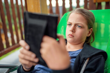 Bored young teen girl with pouting sad expression reading tablet while lounging in green recliner on sunny wooden terrace balcony, authentic child emotion and screen time