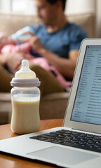 Warm baby bottle with formula milk propped next to a working laptop, and a young father caring and feeding his infant in background. Modern fatherhood concept as balance of parenting and remote work