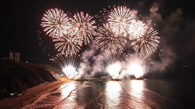 Sweeping high aerial drone shot moving smoothly over a vast dark coastline while simultaneous colorful pyrotechnic displays erupt along the shoreline aerial view, scenic, cinematic