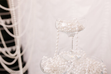 wide glass champagne glasses are arranged in a pyramid and decorated with pearl beads. photo area for a special event and birthday party. glass tableware on a floating light white fabric background.
