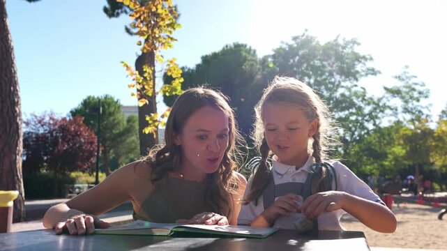Young mother tutoring her elementary school daughter after class. The girl, in her uniform, reads aloud from a book at a park table