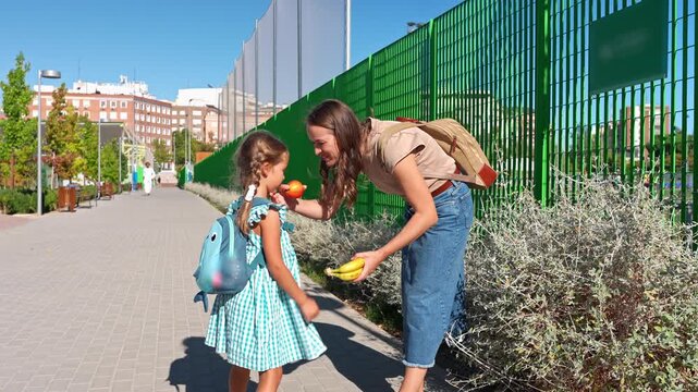 Young mother giving her little girl a healthy fruit snack before she walks to school, then kissing her and waving goodbye on a sunny day