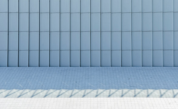 Minimalist blue facade with vertical panels and geometric pavement with sharp shadow.