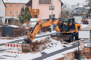 Wheeled excavator digging trench on snowy residential street during winter road construction © Katharina