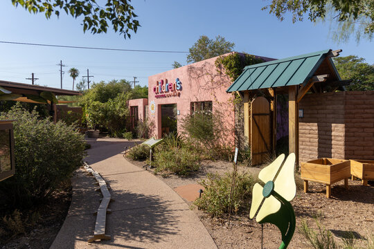 Tucson, Arizona, USA - August 19th, 2025: Colorful pink adobe building entrance to Children's Museum at Tohono Chul Park with green roof wooden gate flower art and desert garden path