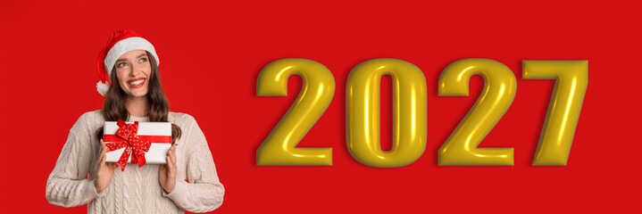 Obraz premium A woman wearing a festive hat smiles while holding a gift in front of a bright red background with the year 2027 displayed. This scene captures a joyful moment during a holiday celebration.