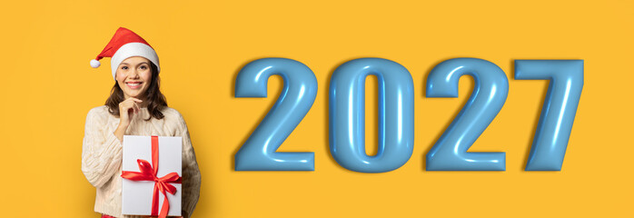 Obraz premium A person wearing a Santa hat holds a gift wrapped with a red ribbon while smiling. The background shows a bright yellow color with large numbers representing the year 2027.