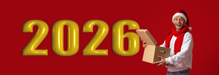 Obraz premium A man wearing a Santa hat and red scarf smiles as he holds a cardboard box in front of large golden numbers indicating the next year. The background is bright red.