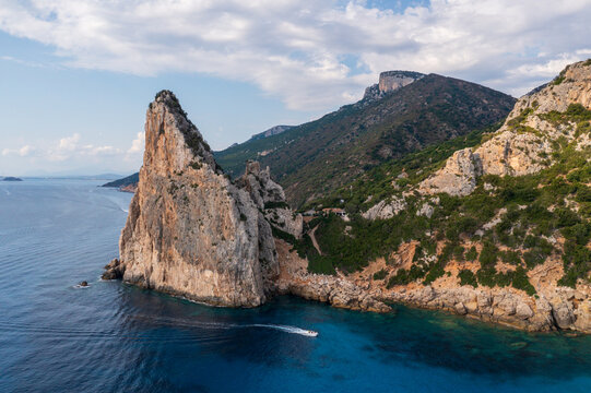 Aerial view of a jagged, imposing rock formation jutting from the azure sea, contrasting with the verdant slopes of the distant mountains, Baunei, Italy.