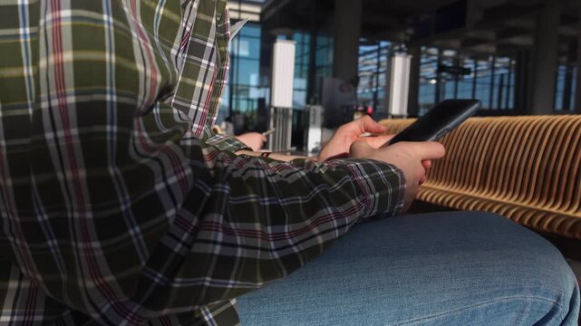Man scrolling phone on bench, plaid shirt and jeans waiting at gate in busy terminal, natural light through large windows, distant flight displays and seating rows, calm focus preparing for connection