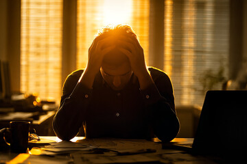 Obraz premium Stressed man with head in hands at a sunset-lit desk, overwhelmed by paperwork and deadlines — office burnout and workplace anxiety scene