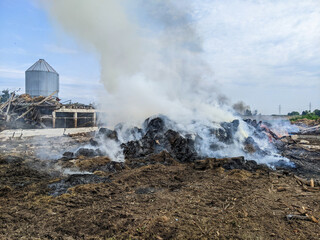 Charred debris and thick smoke blanket a rural farmyard beside a silo, with a ruined barn and scattered wreckage. The scene conveys destruction, danger and the need for cleanup on farmland.