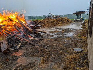 A muddy construction site shows a large pile of wood burning with flames and smoke. A Skid Steer in the background brings wood across the wet ground full of scattered hay and straw on a rural farmyard
