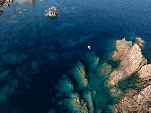 Aerial view of rugged cliffs meet the turquoise sea, a solitary boat adrift in the vastness, where nature's artistry unfolds in a spectacle of raw beauty, Capo Testa, Sardegna, Italy.